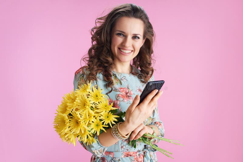 Portrait of happy trendy woman with long wavy brunette hair with yellow chrysanthemums flowers sending text message using smartphone isolated on pink. Portrait of happy trendy woman with long wavy brunette hair with yellow chrysanthemums flowers sending text message using smartphone isolated on pink