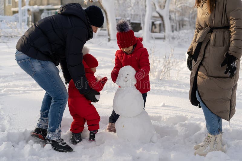 Happy family with two little girls in a winter park on a sunny day sculpts a snowman. Mom, dad and 2 daughters are making snow balls. Winter fun for parents and children. Happy family with two little girls in a winter park on a sunny day sculpts a snowman. Mom, dad and 2 daughters are making snow balls. Winter fun for parents and children.