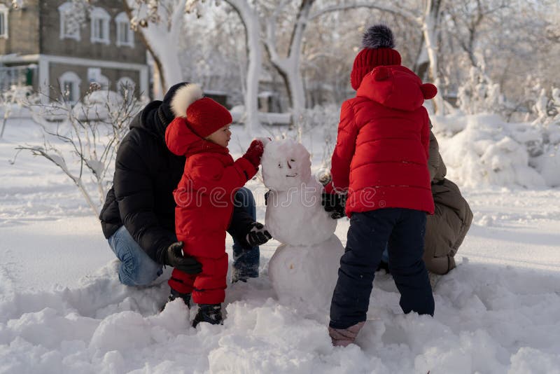 Happy family with two little girls in a winter park on a sunny day sculpts a snowman. Mom, dad and 2 daughters are making snow balls. Winter fun for parents and children. Happy family with two little girls in a winter park on a sunny day sculpts a snowman. Mom, dad and 2 daughters are making snow balls. Winter fun for parents and children.