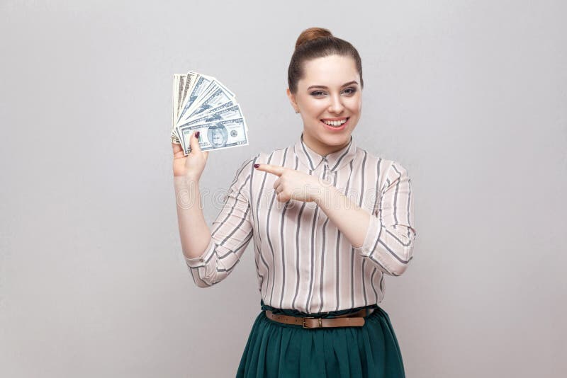 Счастливая удовлетворенная привлекательная молодая коммерсантка в положении striped рубашки, держащ много долларов, указывающ пал стоковые фото Счастливая удовлетворенная привлекательная молодая коммерсантка в положении striped рубашки, держащ много долларов, указывающ пал стоковые фото