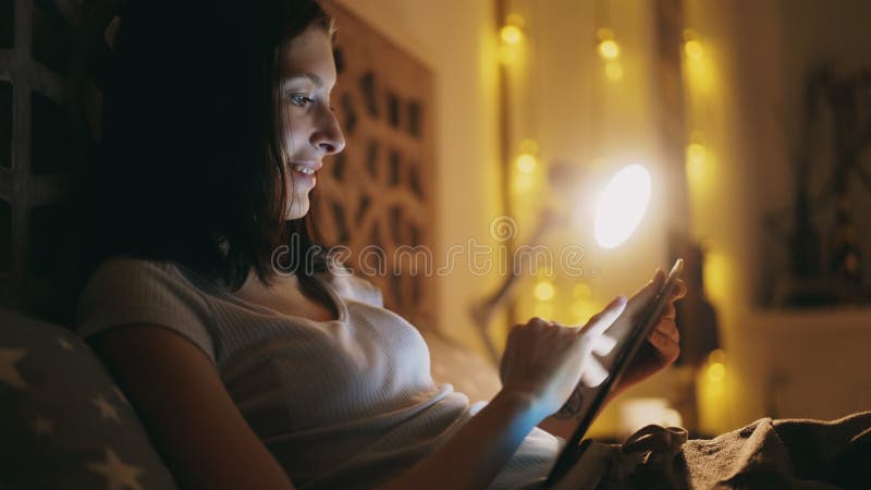 Счастливая усмехаясь женщина используя планшет для делить социальные средства массовой информации лежа в кровати дома на ноче стоковые изображения rf Счастливая усмехаясь женщина используя планшет для делить социальные средства массовой информации лежа в кровати дома на ноче стоковые изображения rf