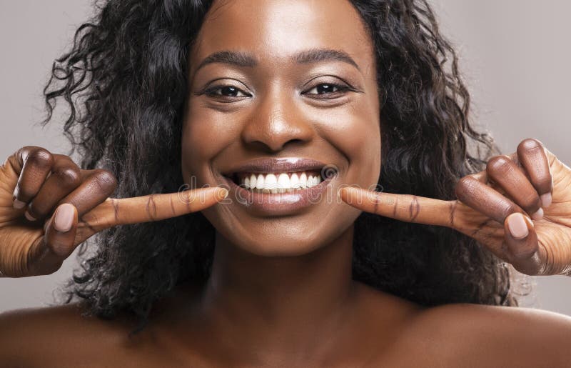 Teeth alignment and whitening concept. Happy african american girl pointing at her wide smile with two forefingers over grey background, closeup. Teeth alignment and whitening concept. Happy african american girl pointing at her wide smile with two forefingers over grey background, closeup