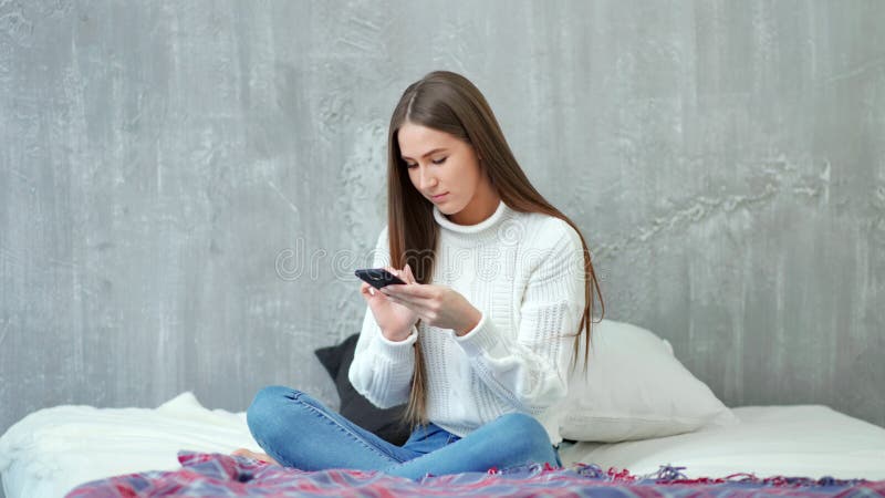 Счастливое молодое европейское сообщение сочинительства женщины используя смартфон сидя на кровати на уютной спальне сток-видео