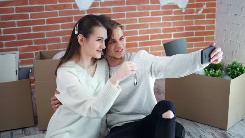 Счастливые молодые пары показывая ключи от новой квартиры принимая selfie используя съемку смартфона среднюю сток-видео
