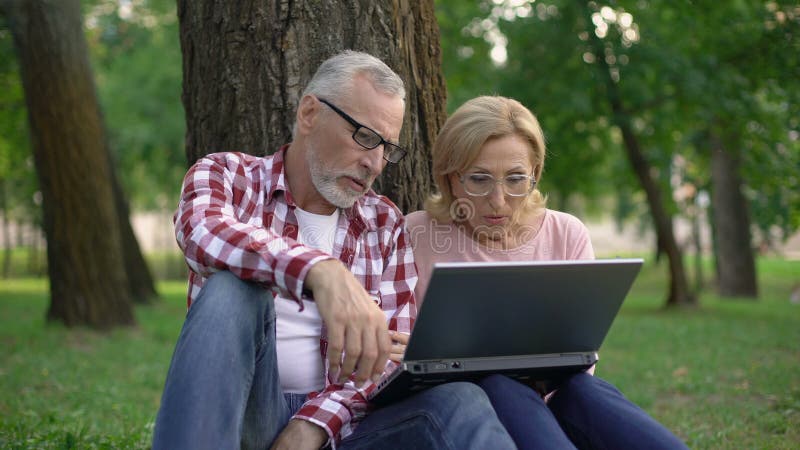 Счастливый выбытые человек и женщина сидя на траве и смотря кино на ПК компьтер-книжки