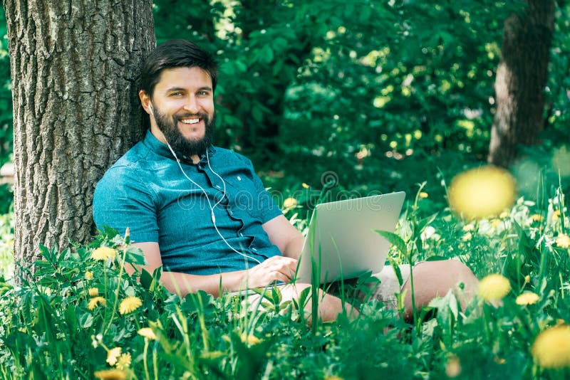 Счастливый жизнерадостный человек битника при компьтер-книжка сидя outdoors на gre стоковые изображения Счастливый жизнерадостный человек битника при компьтер-книжка сидя outdoors на gre стоковые изображения