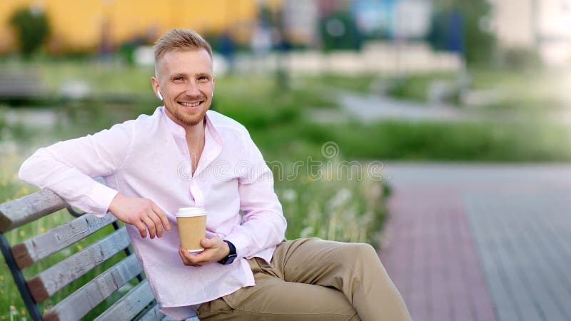 Счастливый случайный парень в беспроводных наушниках сидя на стенде на парке лета держа бумажную кофейную чашку