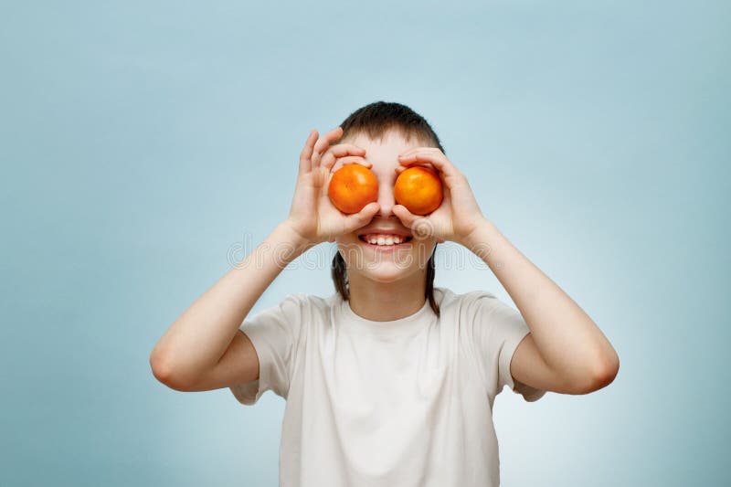 счастливый улыбающийся мальчик с двумя мандаринами, закрывающими глаза на синий фон стоковые фото
