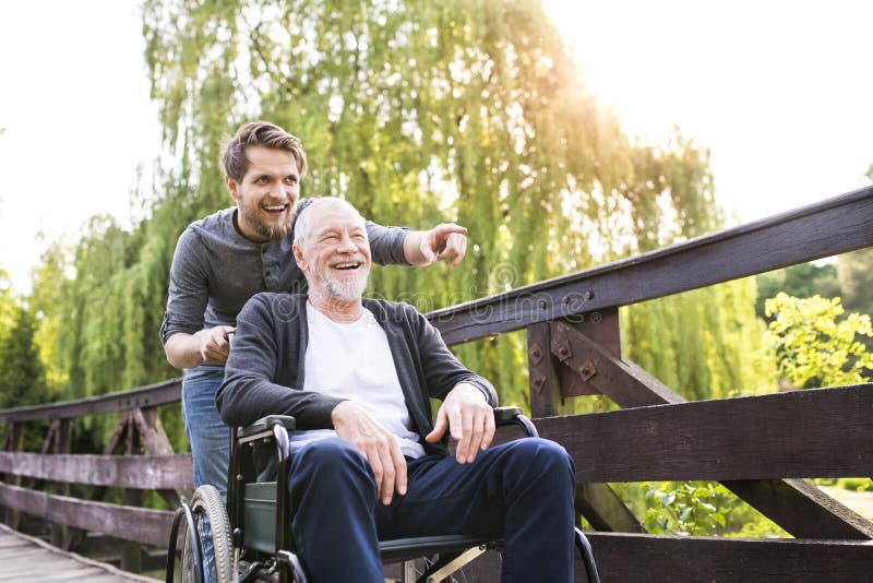 Young hipster son walking with disabled father in wheelchair on wooden bridge at park, pointing finger at something. Carer assisting disabled senior man. Young hipster son walking with disabled father in wheelchair on wooden bridge at park, pointing finger at something. Carer assisting disabled senior man.