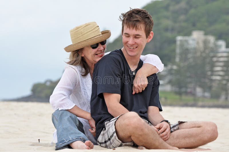 A senior middle-aged mother with her adult son relaxing on a beautiful white sandy beach sharing a joke and laughing with each other. A senior middle-aged mother with her adult son relaxing on a beautiful white sandy beach sharing a joke and laughing with each other.