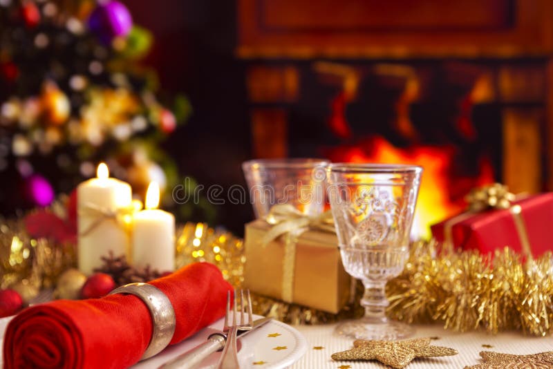 A romantic Christmas dinner table setting with candles and Christmas decorations. A fire is burning in the fireplace and Christmas stockings are hanging on the mantelpiece. A Christmas tree is standing next to the fireplace in the background. A romantic Christmas dinner table setting with candles and Christmas decorations. A fire is burning in the fireplace and Christmas stockings are hanging on the mantelpiece. A Christmas tree is standing next to the fireplace in the background.