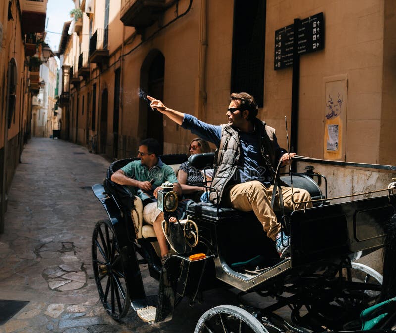 Palma de Mallorca, Spain - May 27, 2016: Extraordinary tour guide in horse carriage buggy points a hand with a cigarette at a landmark for couple of tourists in old town city of Mallorca, Spain. Palma de Mallorca, Spain - May 27, 2016: Extraordinary tour guide in horse carriage buggy points a hand with a cigarette at a landmark for couple of tourists in old town city of Mallorca, Spain