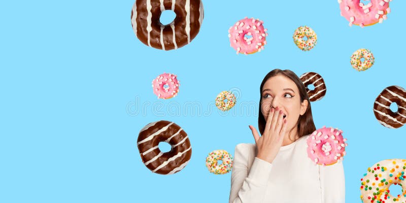 Удивительная молодая женщина, покрывающая рот, а разноцветные пончики плывут вокруг нее на ярко-синем фоне. стоковое фото