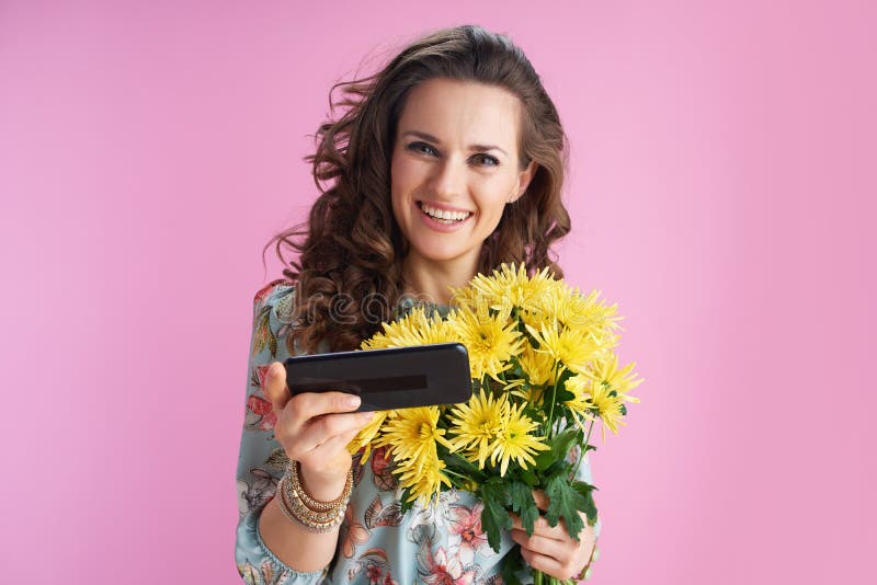 Portrait of smiling modern 40 years old woman in floral dress with yellow chrysanthemums flowers sending text message using smartphone isolated on pink background. Portrait of smiling modern 40 years old woman in floral dress with yellow chrysanthemums flowers sending text message using smartphone isolated on pink background