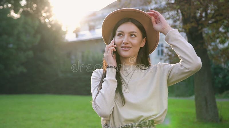 Улыбающаяся молодая женщина в бейдж-шляпе, стоящая рядом с зеленым деревом во дворе дома и разговаривающая по телефону
