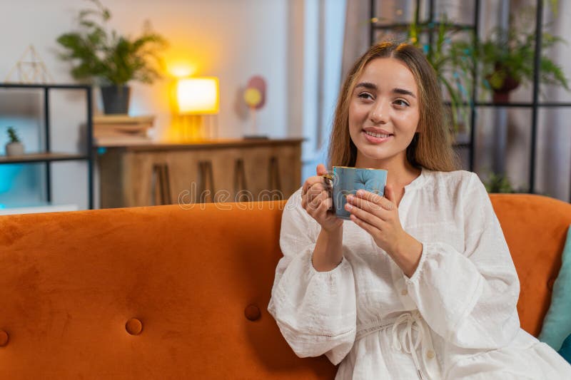 Улыбающаяся молодая женщина, сидя на диване, пьет чашку теплого кофе или травяной чай утром дома стоковые фотографии rf