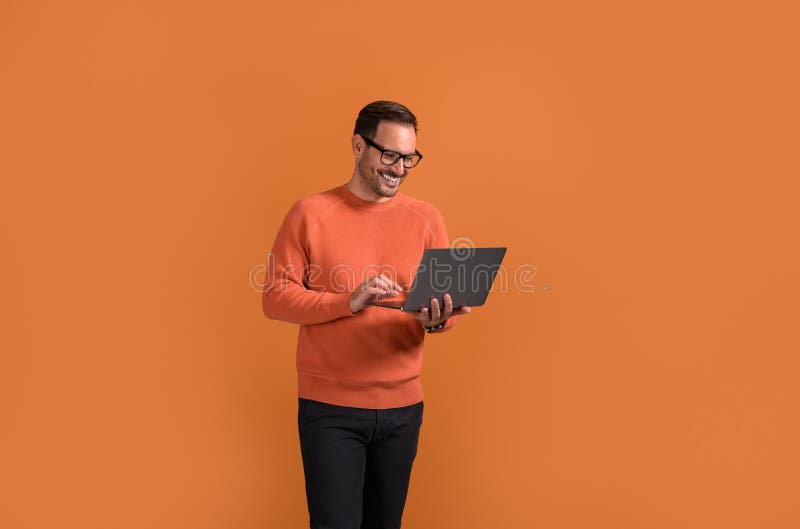 Smiling male manager checking e-mails over laptop while standing confidently on orange background. Smiling male manager checking e-mails over laptop while standing confidently on orange background.