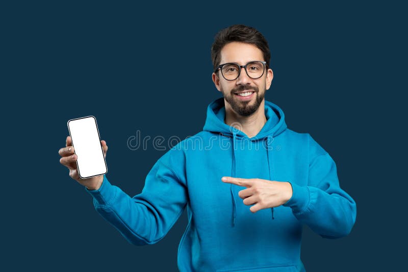 A cheerful young man with a beard and glasses is wearing a blue hoodie and presenting a smartphone with its screen facing forward towards the viewer. He is pointing at the screen. A cheerful young man with a beard and glasses is wearing a blue hoodie and presenting a smartphone with its screen facing forward towards the viewer. He is pointing at the screen