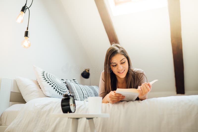 Улыбаясь книги и чтения молодой женщины в тепле кофе в солнечном apartmentrelaxation в теплом homehygge bedsunny light стоковые изображения