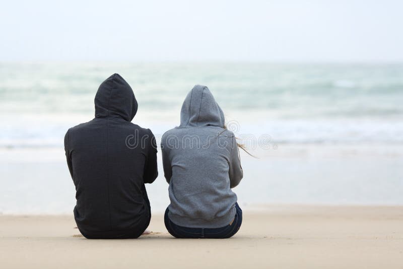 Back view of two sad teenagers sitting on the sand of the beach and looking at horizon in a bad weather day. Back view of two sad teenagers sitting on the sand of the beach and looking at horizon in a bad weather day