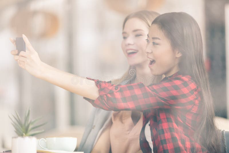 Усмехаясь молодые женщины используя смартфон пока выпивающ кофе совместно стоковые фотографии rf Усмехаясь молодые женщины используя смартфон пока выпивающ кофе совместно стоковые фотографии rf