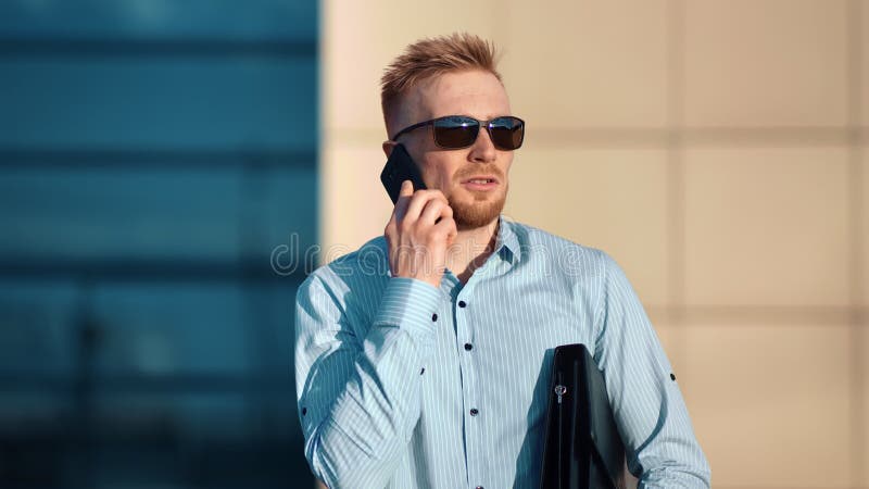 Усмехаясь мужской бизнесмен в солнечных очках говоря используя съемку портфеля удерживания смартфона среднюю