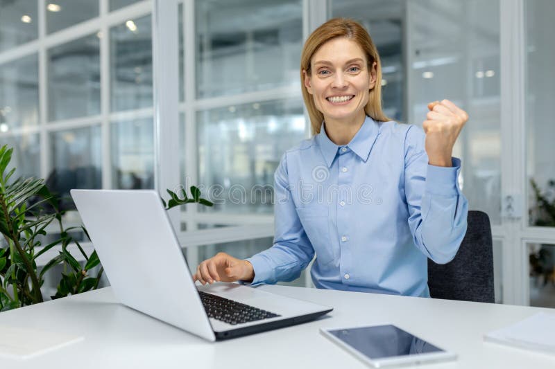 A professional businesswoman in a blue shirt celebrates success while working on a laptop in a modern office environment. She shows joy and satisfaction. A professional businesswoman in a blue shirt celebrates success while working on a laptop in a modern office environment. She shows joy and satisfaction.