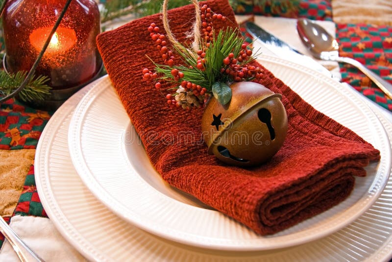 This is a shot of a Christmas place setting with ivory plate and bowl, red fabric napkin, bell Christmas ornament and a red lit bubble glass lantern. The plate is sitting on a country Christmas print quilted tablecloth. There is a fir tree branch decorating the table. This is a shot of a Christmas place setting with ivory plate and bowl, red fabric napkin, bell Christmas ornament and a red lit bubble glass lantern. The plate is sitting on a country Christmas print quilted tablecloth. There is a fir tree branch decorating the table.