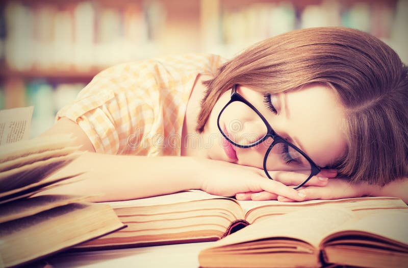Утомленная девушка студента с стеклами спать на книгах в библиотеке стоковые изображения Утомленная девушка студента с стеклами спать на книгах в библиотеке стоковые изображения