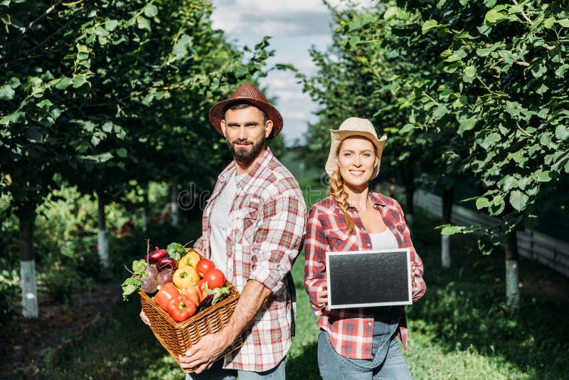 Farmers holding fresh organic vegetables in basket and blank chalkboard while smiling at camera. Farmers holding fresh organic vegetables in basket and blank chalkboard while smiling at camera