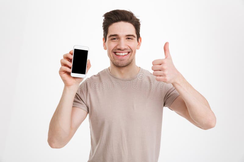 Photo of brunette guy in beige t-shirt holding cell phone with copyspace black screen and gesturing thumb up isolated over white wall. Photo of brunette guy in beige t-shirt holding cell phone with copyspace black screen and gesturing thumb up isolated over white wall
