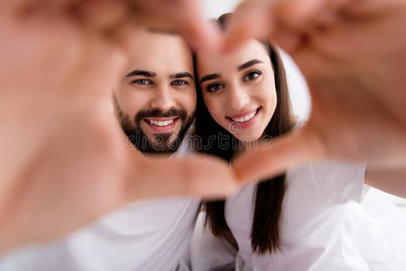 Photo of pretty adorable couple wear white t-shirts embracing showing arms heart indoors apartment bedroom. Photo of pretty adorable couple wear white t-shirts embracing showing arms heart indoors apartment bedroom.