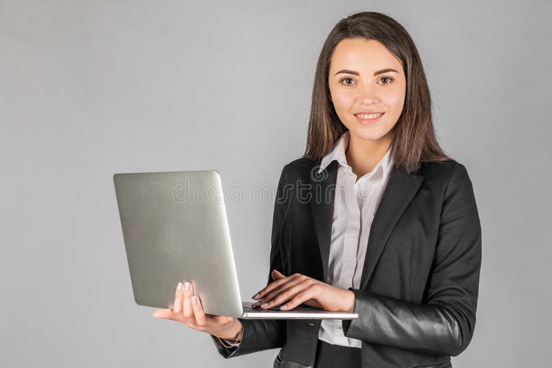 Фотография молодой радостной деловой женщины, стоящей над серой стеной с ноутбуком