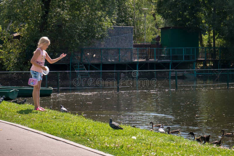 MOSCOW - AUGUST 2, 2018: Blond teenage girl feeding bread to pigeons and ducks swimming in pond in park on summer day. MOSCOW - AUGUST 2, 2018: Blond teenage girl feeding bread to pigeons and ducks swimming in pond in park on summer day