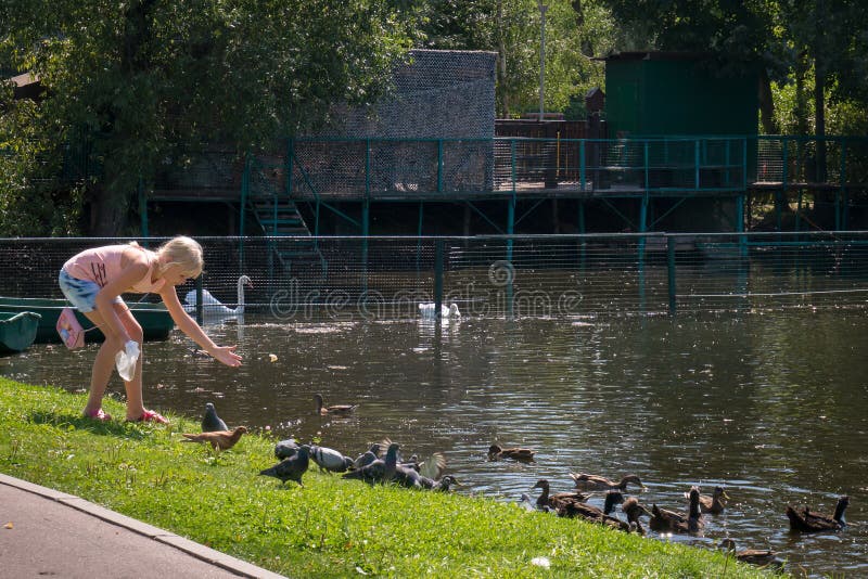 MOSCOW - AUGUST 2, 2018: Blond teenage girl feeding bread to pigeons and ducks swimming in pond in park on summer day. MOSCOW - AUGUST 2, 2018: Blond teenage girl feeding bread to pigeons and ducks swimming in pond in park on summer day