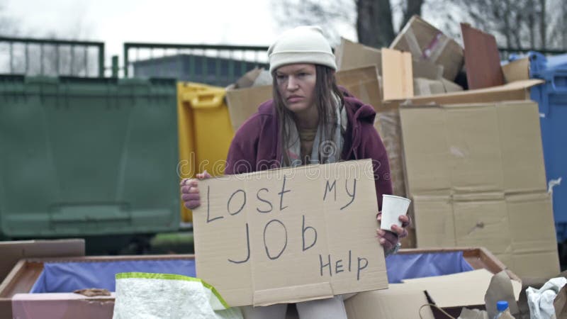 Холодная, незабываемая молодая женщина сидит с попрошайкой чашкой за кучей мусора и держит рукописного писка потерянной работой