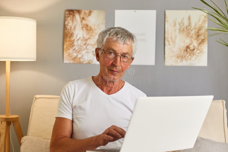Positive grey-haired senior man in casual white T-shirt and glasses sitting on couch at home in living room interior using laptop answering e-mails typing massage browsing internet. Positive grey-haired senior man in casual white T-shirt and glasses sitting on couch at home in living room interior using laptop answering e-mails typing massage browsing internet.