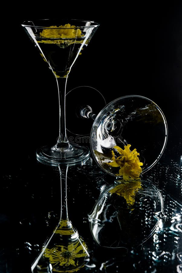 Martini glasses containing water with yellow flowers in them. One glass is standing and the other one is fallen, on a black background. Martini glasses containing water with yellow flowers in them. One glass is standing and the other one is fallen, on a black background.