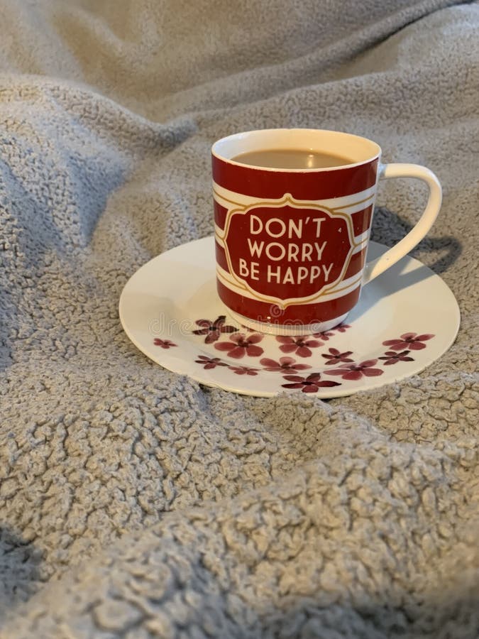 Cup of coffee with text "Don't worry be happy" on white saucer with red pink flowers on bed. Cup of coffee with text "Don't worry be happy" on white saucer with red pink flowers on bed