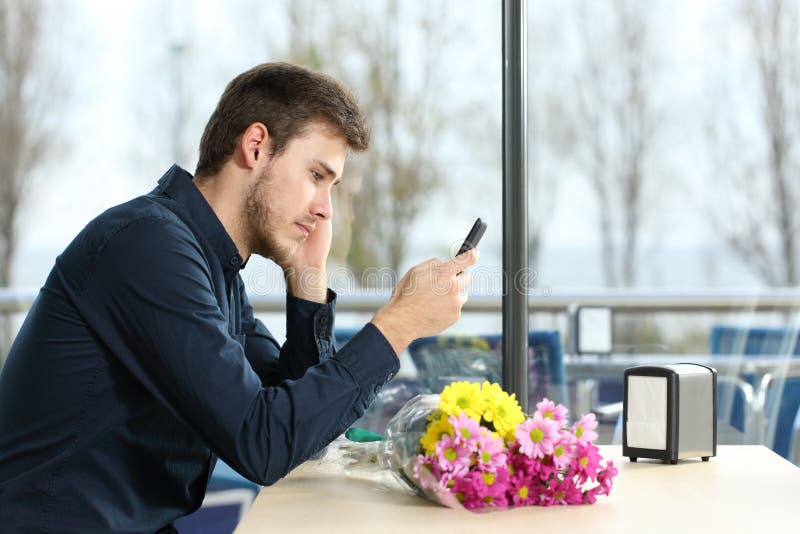 Sad man with a bouquet of flowers stood up in a date checking phone messages in a coffee shop. Sad man with a bouquet of flowers stood up in a date checking phone messages in a coffee shop