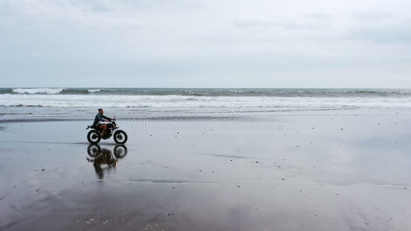 Человек в ехать мотоцикле на пляже винтажный мотоцикл на заходе солнца пляжа на Бали Молодой мужчина хипстера наслаждаясь свободо