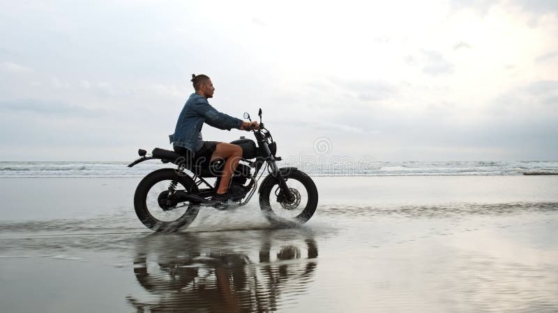 Человек в ехать мотоцикле на пляже винтажный мотоцикл на заходе солнца пляжа на Бали Молодой мужчина хипстера наслаждаясь свободо