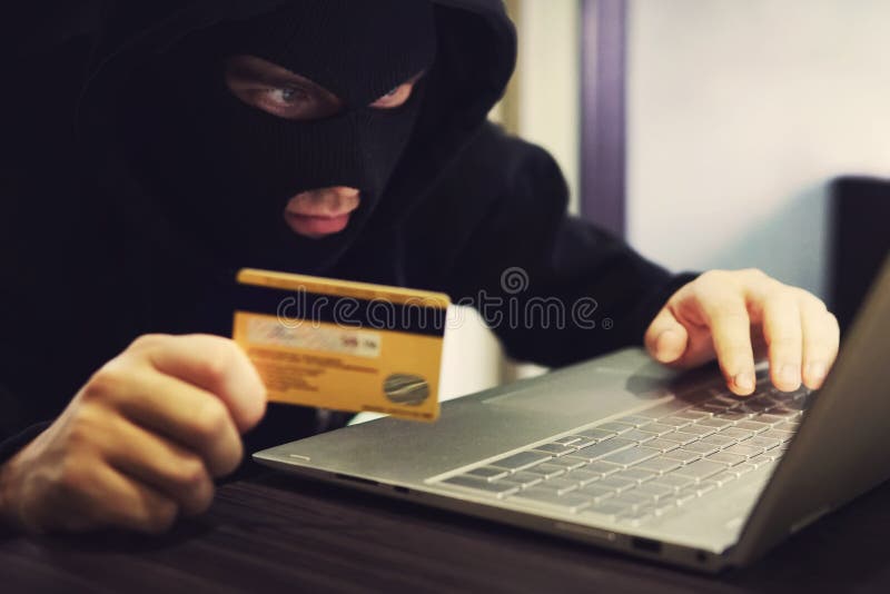 Man in robber mask and hood misappropriates personal bank data. Cyber fraudster attacks online banking system. Hacker makes financial fraud via internet. Digital crime with the use of payment card. Man in robber mask and hood misappropriates personal bank data. Cyber fraudster attacks online banking system. Hacker makes financial fraud via internet. Digital crime with the use of payment card