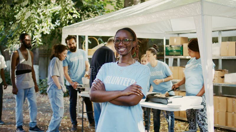 Portrait of african american lady wearing glasses outside with arms crossed and eyes fixed on camera. At food drive, volunteers are aiding the poor, needy and homeless people. Zoom-out, handheld. Portrait of african american lady wearing glasses outside with arms crossed and eyes fixed on camera. At food drive, volunteers are aiding the poor, needy and homeless people. Zoom-out, handheld.