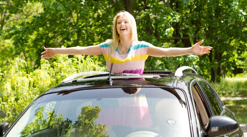 Playful vivacious young blond woman standing in a car sunroof with her arms outstretched laughing happily in the sunshine. Playful vivacious young blond woman standing in a car sunroof with her arms outstretched laughing happily in the sunshine