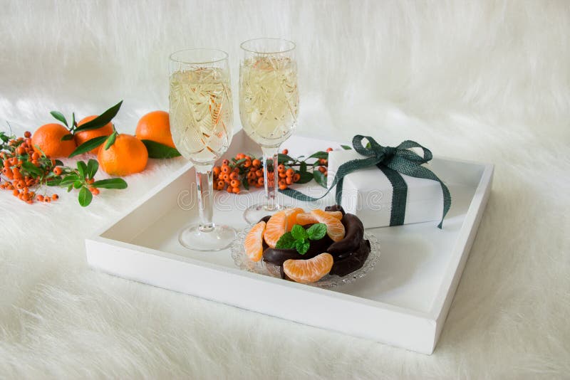 Tray with champagne in 2 crystal glasses, tangerines slices, chocolate and gift on background of rowan twigs, tangerines all on an artificial white fur. Champagne, chocolate, tangerines. Horizontal. Tray with champagne in 2 crystal glasses, tangerines slices, chocolate and gift on background of rowan twigs, tangerines all on an artificial white fur. Champagne, chocolate, tangerines. Horizontal.