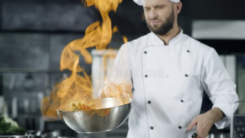 Шеф-повар человека варя в лотке с огнем в замедленном движении на кухне Молодой шеф-повар