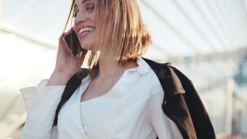 Шикарная молодая женщина в стильной белой блузке счастливо говоря на ее телефоне, дает красивую улыбку, смеется над Бизнес