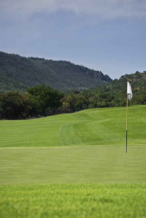 Flagpole is positioned to the right of the frame, behind that the fairway approach of one of the holes, showing also the thick rough bush area that surrounds all the holes of the Sun City Gary Player Golf Course. Flagpole is positioned to the right of the frame, behind that the fairway approach of one of the holes, showing also the thick rough bush area that surrounds all the holes of the Sun City Gary Player Golf Course.