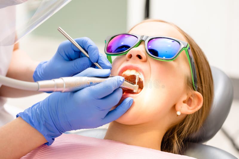 Dental hygienist performing teeth cleaning procedure to elementary age girl in pediatric dental clinic. Doctor using prophy brush on handpiece to remove hardened deposits of mineralized plaque or tartar. Dental hygienist performing teeth cleaning procedure to elementary age girl in pediatric dental clinic. Doctor using prophy brush on handpiece to remove hardened deposits of mineralized plaque or tartar